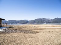 a dirt covered field in front of mountains and a man fishing on the lakefront