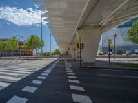 a car sitting at an empty street underneath a overpass overhang that contains a train stop, and cars and a pedestrian