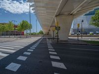 a car sitting at an empty street underneath a overpass overhang that contains a train stop, and cars and a pedestrian