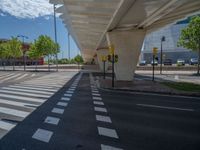 a car sitting at an empty street underneath a overpass overhang that contains a train stop, and cars and a pedestrian