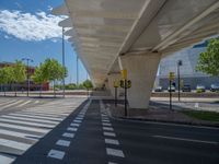 a car sitting at an empty street underneath a overpass overhang that contains a train stop, and cars and a pedestrian