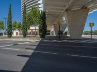 a car sitting at an empty street underneath a overpass overhang that contains a train stop, and cars and a pedestrian