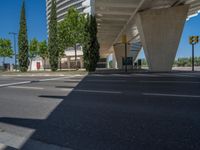 a car sitting at an empty street underneath a overpass overhang that contains a train stop, and cars and a pedestrian