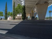 a car sitting at an empty street underneath a overpass overhang that contains a train stop, and cars and a pedestrian