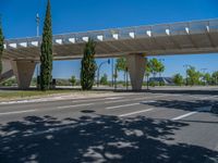 a car sitting at an empty street underneath a overpass overhang that contains a train stop, and cars and a pedestrian