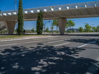 a car sitting at an empty street underneath a overpass overhang that contains a train stop, and cars and a pedestrian