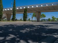 a car sitting at an empty street underneath a overpass overhang that contains a train stop, and cars and a pedestrian