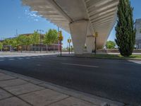 a car sitting at an empty street underneath a overpass overhang that contains a train stop, and cars and a pedestrian