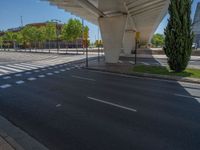 a car sitting at an empty street underneath a overpass overhang that contains a train stop, and cars and a pedestrian